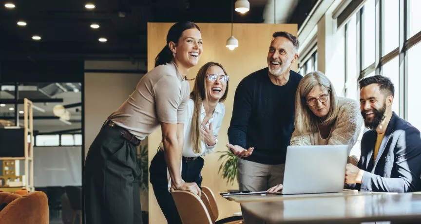 Pět kolegů různého věku se společně směje u laptopu v moderní kanceláři – ilustrace pozitivní firemní kultury a employer brandingu
