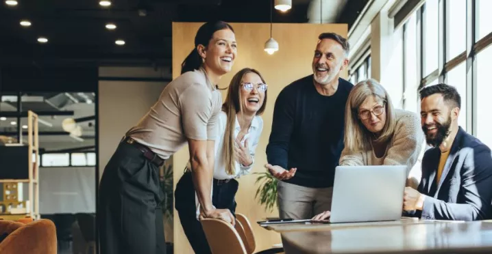 Pět kolegů různého věku se společně směje u laptopu v moderní kanceláři – ilustrace pozitivní firemní kultury a employer brandingu